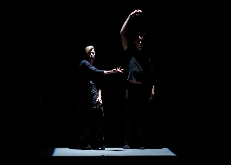 The two dancers dressed in black are in a square of dim light and  strike vogue-like poses